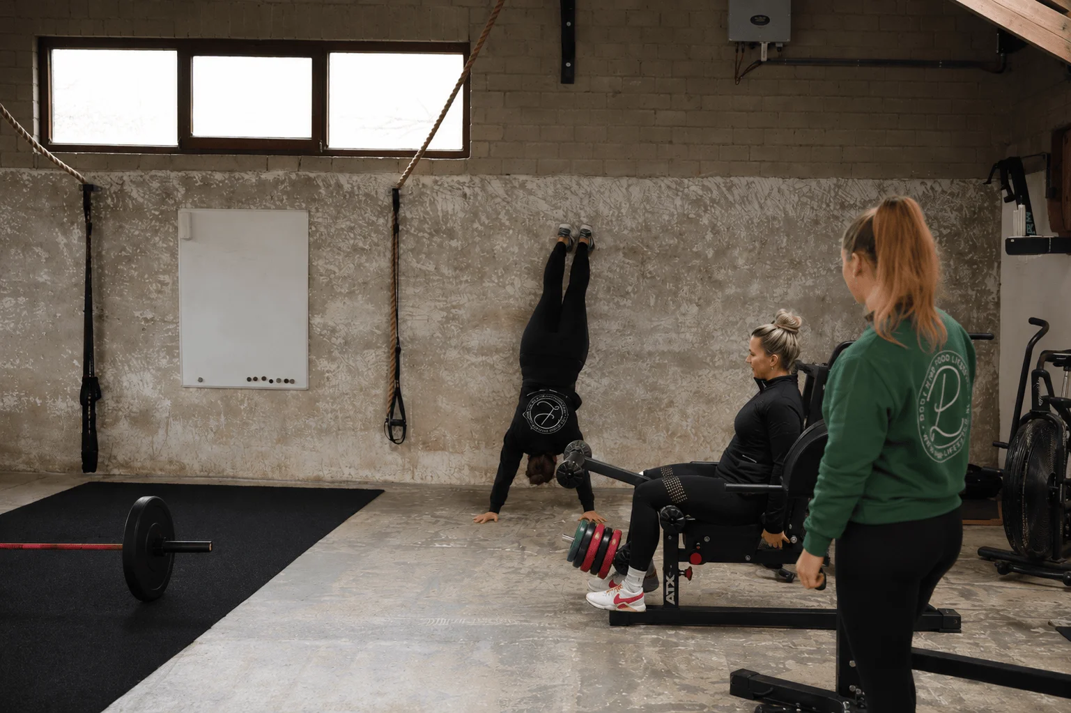 Drie vrouwen doen verschillende oefeningen tegelijk — handstand, leg-press en airbike